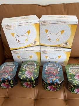 Four Pokémon "Elite Trainer Box" boxes and four floral-patterned tins, including what appear to be "Ascended Heroes" tins, are stacked on a brown leather couch. The boxes feature Pikachu and display "30" anniversary branding, while the post highlights the user's excitement at finding the "Ascended tins" available on the shelf.