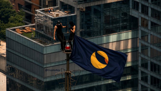 Two climbers plant a large LUNC (Terra Classic) flag atop a city rooftop, symbolizing the community's bold ascent into the mainstream and reflecting the post's message that LUNC has moved beyond its early days to become a recognized force in the crypto skyline. The watermark "@CryptoAnu" is visible in the lower left of the image.