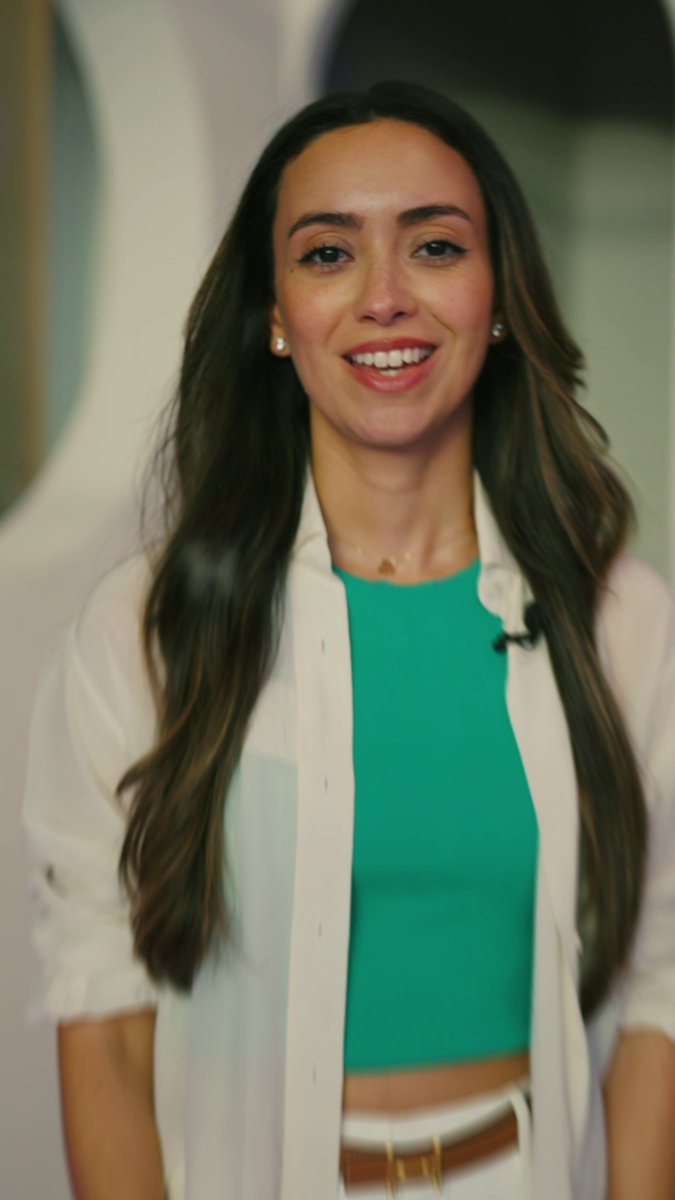 In the image, a woman wearing a white shirt and green top stands against a gray background. She has long brown hair that falls over her shoulders. The woman appears to be smiling at the camera, possibly posing for a photo or participating in an event related to blockchain technology.