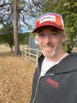 A bearded man wearing a colorful trucker hat and a black 'Everglades Towing Club' hoodie, taking a selfie outdoors near trees and a fence.