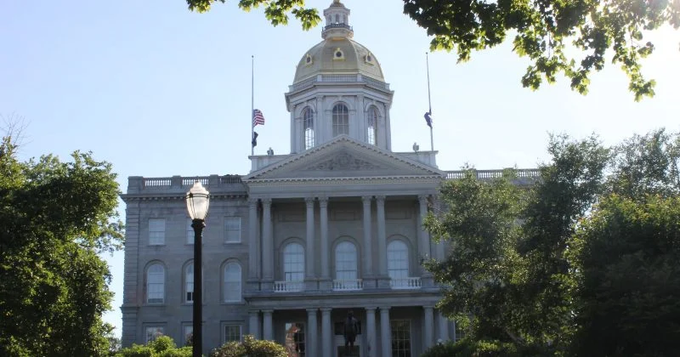 A photograph of a historic government building featuring a gold-domed cupola, American and state flags, classical architecture with columns, framed by green trees and an ornate street lamp.