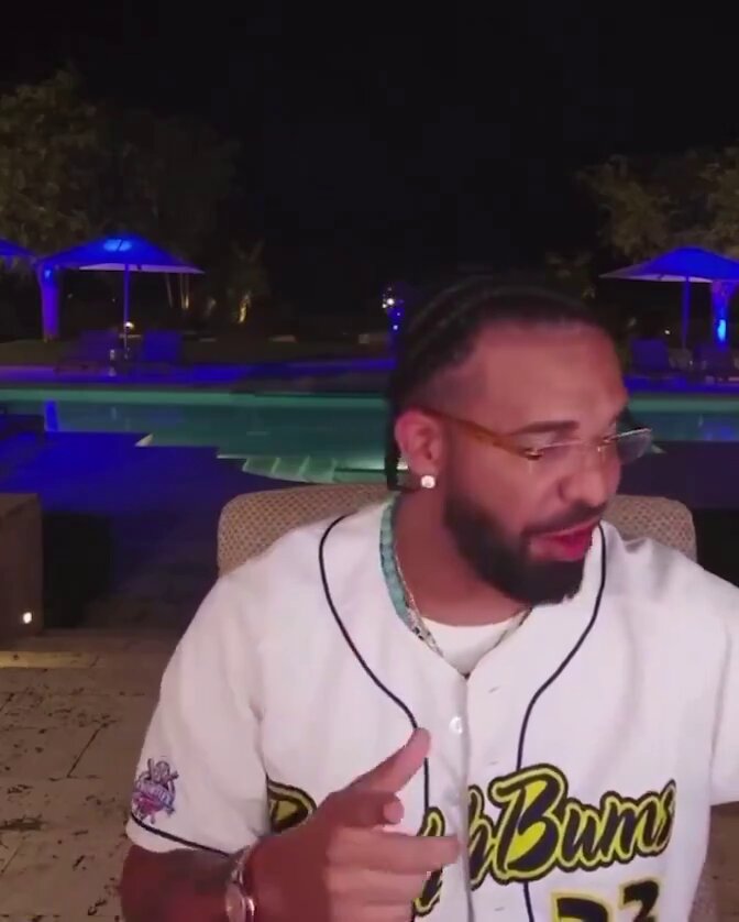 The image features a man wearing a baseball jersey with the number 33, sitting at an outdoor table near a pool area. He has a beard and is wearing glasses. The background includes several umbrellas and potted plants, creating a relaxed atmosphere around him. The caption of the image reads "Bro worth half a bill and still more grateful than some of you Big W for @wallchain new system Small creators should definitely take those deals!".