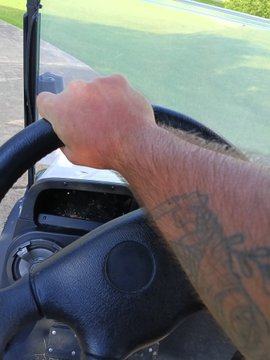 A first-person view of a person's tattooed arm gripping the steering wheel of a golf cart on a grassy course.