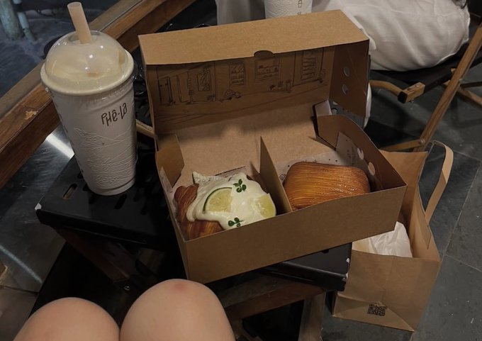 An iced drink labeled "PHE-12" sits next to an open cardboard box containing pastries and a croissant.