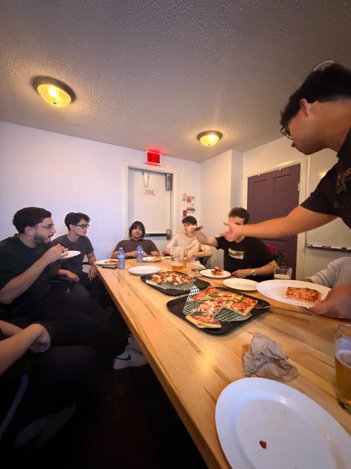 A casual photo of a group of young men eating pizza together around a long wooden table in a brightly lit room.
