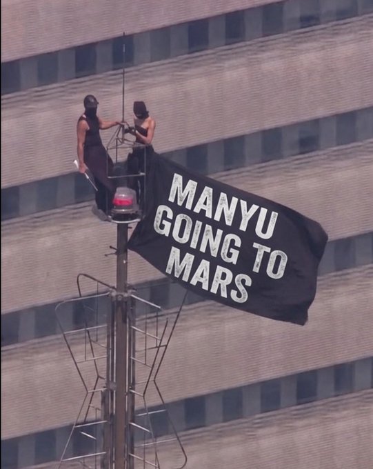 Same image as Image 2: two individuals on a tower holding a black 'MANYU GOING TO MARS' banner.