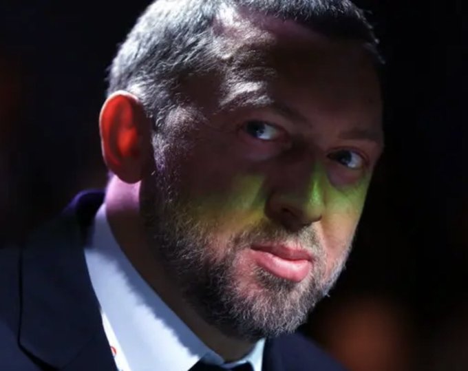 A close-up photograph of a bearded man in a suit looking to the side with a slight smirk, lit with dramatic lighting.