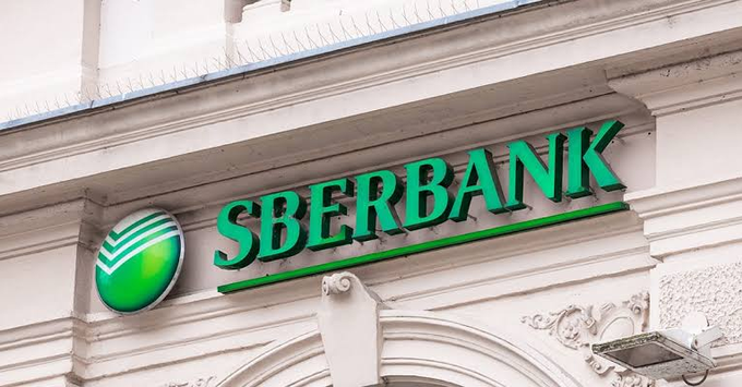 The image shows the Sberbank logo and signage mounted on a building facade under a blue sky, illustrating the post's announcement that Sberbank, Russia's largest bank, will begin offering crypto-backed loans to clients starting in September.
