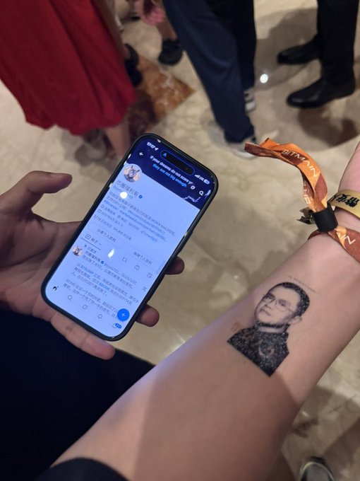 A person holds up a smartphone displaying a social media feed while showing off a wrist tattoo of Binance founder Changpeng Zhao ("CZ") at an event, accompanied by orange wristbands. The image humorously captures the unexpected encounter, showing CZ's likeness inked on the attendee's arm as referenced in the post.