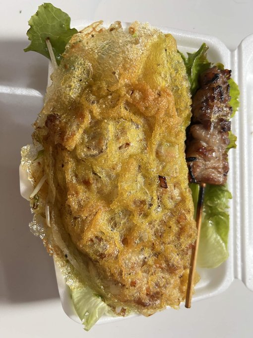 A close-up of an Asian-style dish in a white styrofoam takeout container, featuring a folded egg omelet over rice with bean sprouts, served with grilled meat skewers on a bed of lettuce.