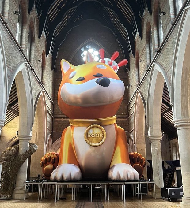 A giant inflatable orange Shiba Inu dog wearing a gold "BONK" tag on its collar, displayed inside a building with Gothic-style stone arches.