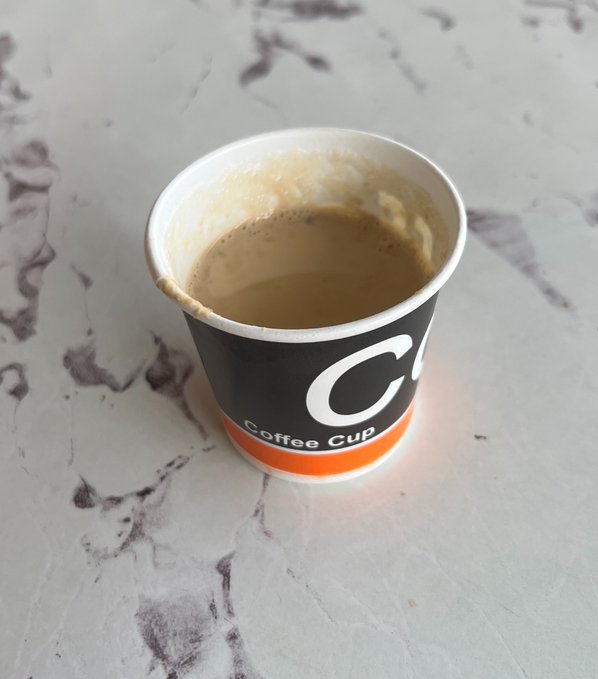 A black paper coffee cup with an orange stripe and the text 'Coffee Cup' sits on a white marble surface, partially filled with coffee.