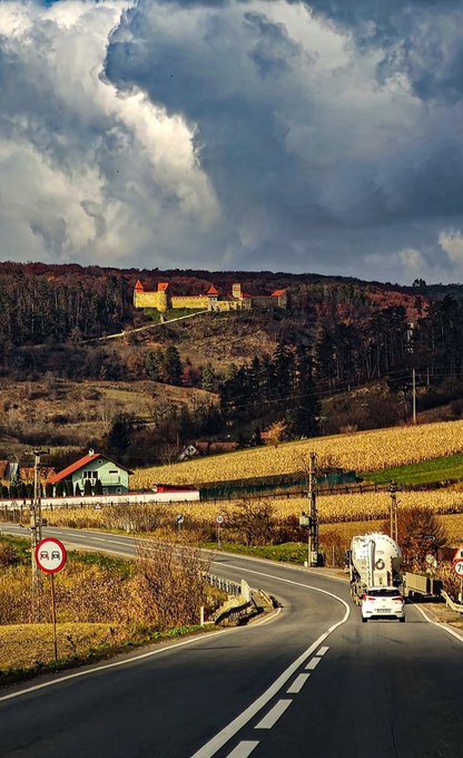 A winding country road cuts through a rolling autumn landscape beneath dramatic cloudy skies, with a white tanker truck and a silver car traveling along it toward a small hillside village. A road sign with a red circle and diagonal line (no entry) is visible in the foreground, while golden cornfields and forested hills with scattered red-roofed houses complete the Eastern European rural scene—fitting for a post commemorating day 661 of the $ENA countdown.
