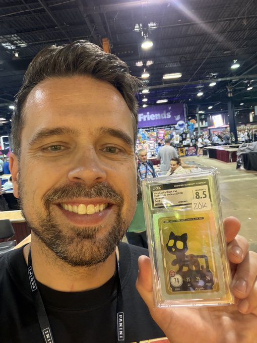 A smiling man at a trading card convention holds up a CGC graded 8.5 copy of the Venifuumi Chrome Sapphire 2024 Pokémon card with a black cat illustration, labeled "ZOK".