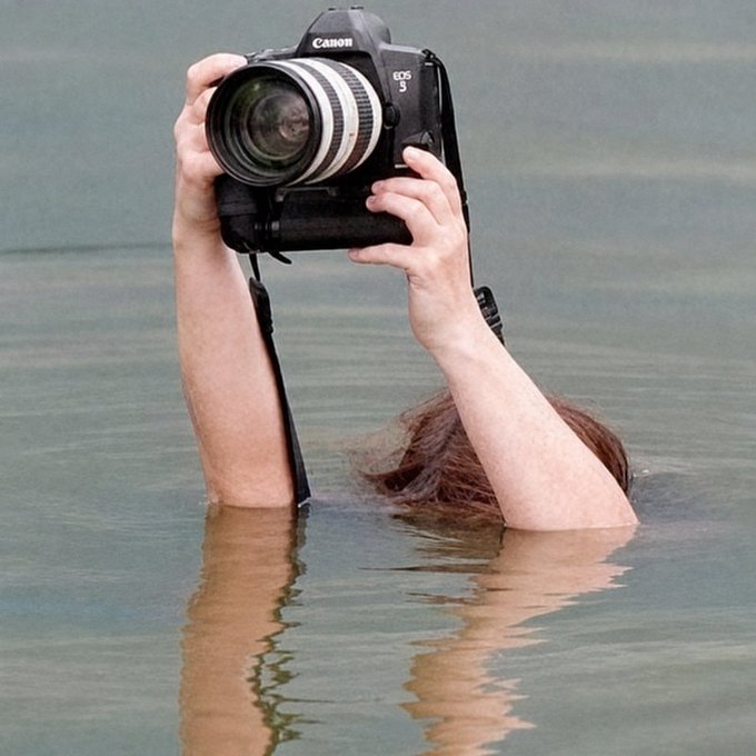 A photographer stands chest-deep in water while raising a Canon EOS 5 camera to capture an unusual angle, humorously illustrating an “intern” going to any length for the shot.