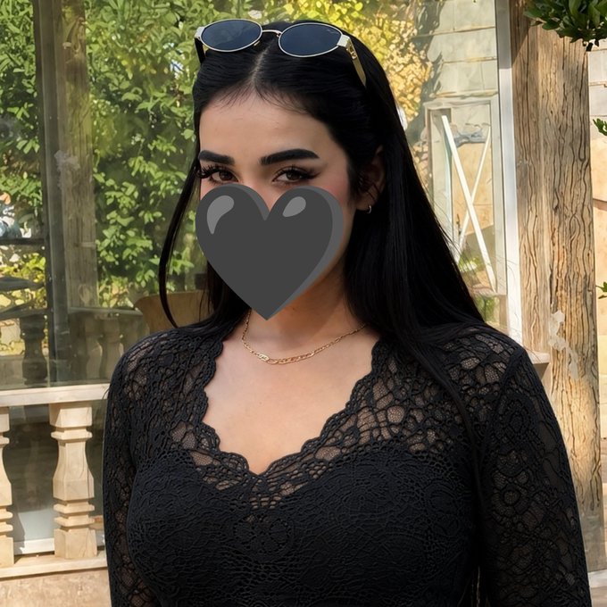 A woman with long dark hair, sunglasses on her head, and a delicate gold necklace stands outdoors in a black lace top, her face partially covered by a heart graphic.