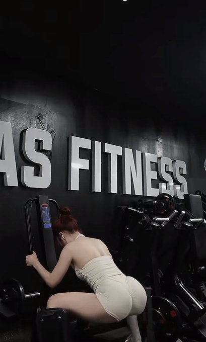 Photo of a woman with red hair in a beige outfit working out on gym equipment in front of a wall displaying large 'AS FITNESS' lettering.