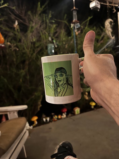 A hand gives a thumbs-up while holding a white coffee mug featuring a green illustrated portrait of a person with glasses, set against an outdoor garden scene at night.