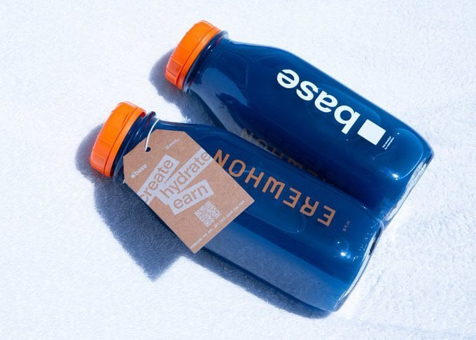Two blue Baes water bottles with orange caps resting on white textured fabric, displaying the tagline 'CREATE, HYDRATE, EARN' and a tag reading 'EREWON'.