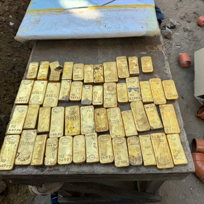 Numerous gold bars stamped with serial numbers and 'NI' markings are arranged on a weathered wooden table, with a notepad resting behind.