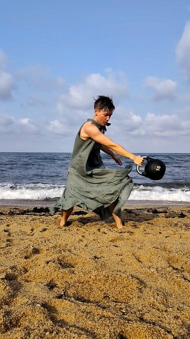 A person in a flowing gray-green dress stands on a sandy beach, holding a black helmet labeled 'No.' against a backdrop of blue ocean and cloudy sky.