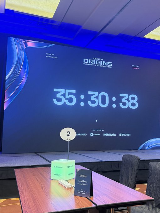 A conference screen displays the 'ORIGINS HACKATHON' logo with a countdown timer reading '35:30:38', supported by Gardana, NOWNodes, and SOLANA, with a table number '2' in the foreground.