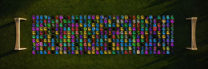 An aerial view of a large rectangular arrangement of many colorful small toy cars organized in neat rows on grass, flanked by two small structures.