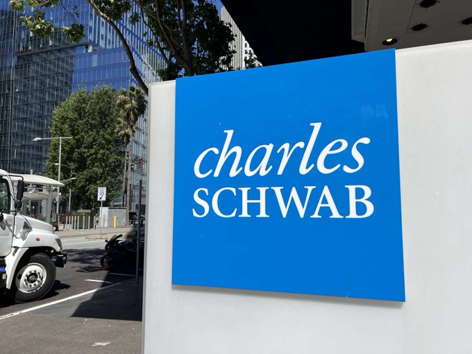 A blue and white 'Charles Schwab' company sign displayed outdoors with a truck and office building visible in the background.