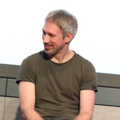 The image shows a man with light brown hair and a beard, wearing an olive green t-shirt, appearing to be seated at what looks like a panel discussion or interview setting. The accompanying post references Gavin Wood (@gavofyork), co-founder of Ethereum and founder of Polkadot ($DOT), suggesting the image depicts him during a public speaking engagement where he is likely discussing blockchain technology and Polkadot's market position.