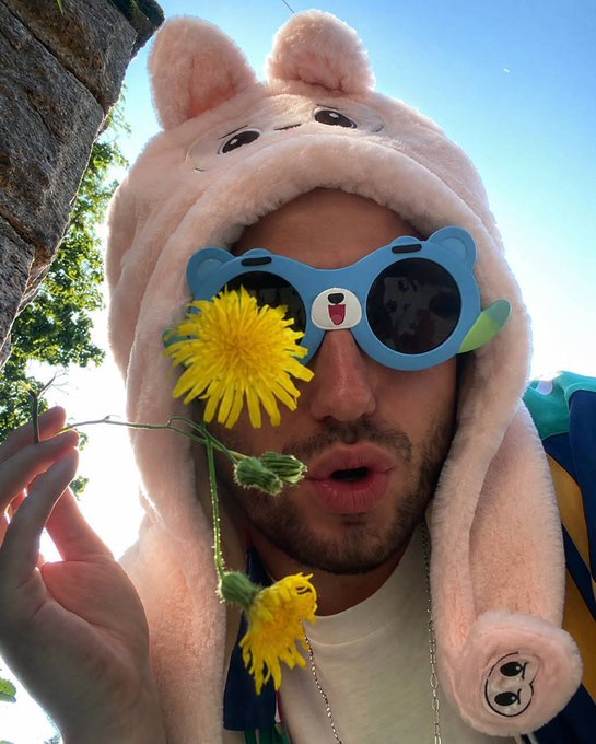A man wearing a pink bear-themed plush hat and blue bear-shaped sunglasses holds a yellow dandelion flower up to his face.