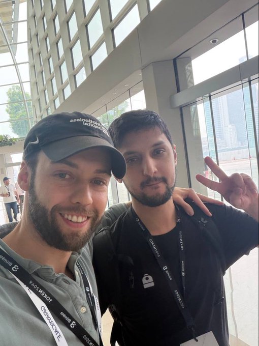 A selfie of two smiling men, one wearing a black cap with 'Decentralized Industries' text and another making a peace sign, both with 'TOKEN2049' lanyards visible.