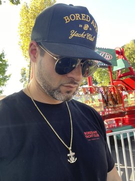 A man wearing a navy "BORED Yacht Club" cap, aviator sunglasses, a Chanel pendant necklace, and a black "General Tso's Club" t-shirt poses at what appears to be an outdoor event.