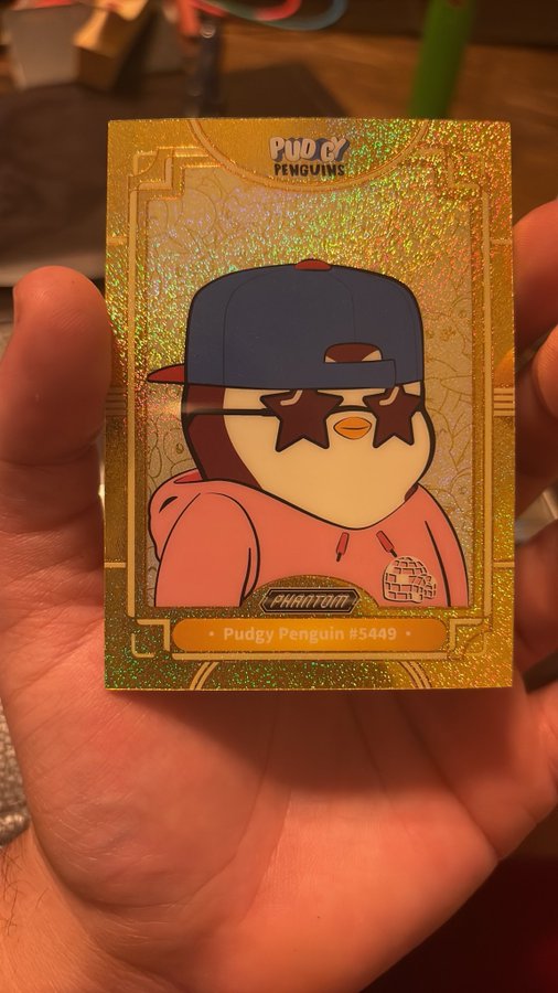 A glowing gold "Pudgy Penguins #5449" collectible card is held up in hand, featuring the signature star-shaped sunglasses penguin wearing a pink hoodie and a blue cap. The accompanying post celebrates the rare gold tier pull from the Pudgy Penguins collection by Web3 Attorneys, with the "Pharaoh" label and gold holographic design marking it as a premium find.