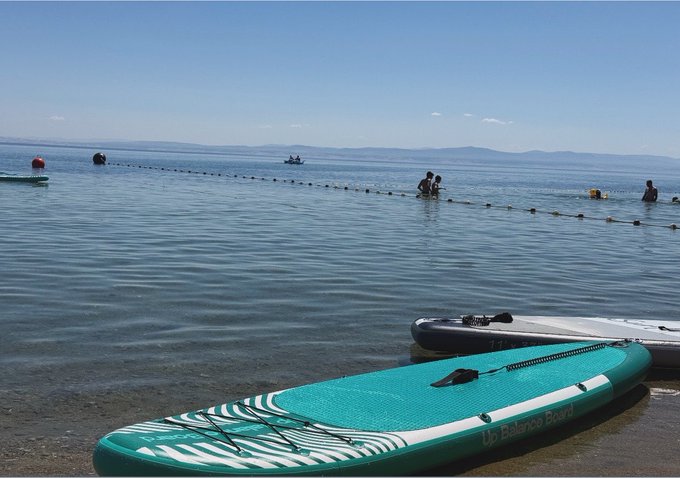 A teal and white "Up Balance Board" paddleboard lying on the shore of a lake or sea where swimmers and boaters can be seen in the distance under a clear blue sky.