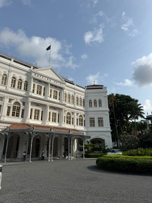 The image shows the iconic white facade of the Raffles Hotel in Singapore, with its name clearly displayed above the columned entrance and a flag flying on top. This grand colonial-era building serves as the venue for "Re-DeFining Finance," an event hosted by Fluid and @agi3official that brought together Mastercard, @FTDA_US, @GSR_io, @ethereuminsti, and @JupiterExchang to discuss the future of finance.