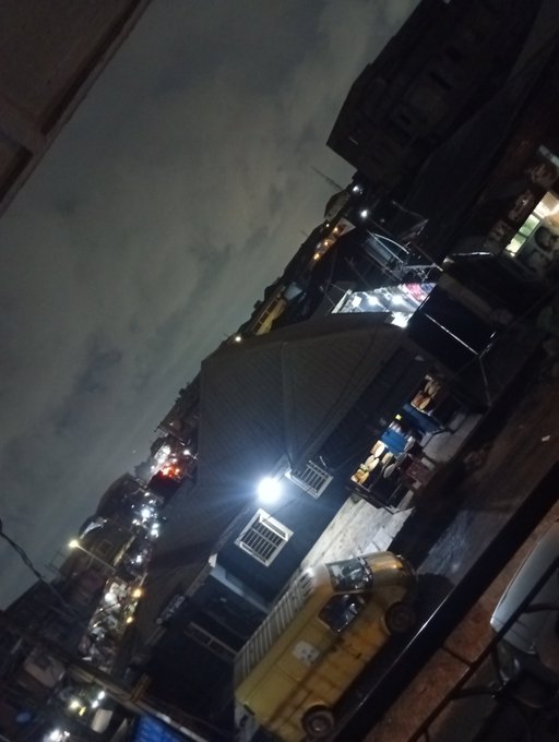 A nighttime cityscape photo taken at a tilted angle shows illuminated street vendors, a yellow vehicle, and a building with a bright overhead light against a cloudy sky.