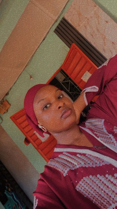 A vertical selfie of a woman wearing a pink headwrap and a red and white beaded traditional outfit, posed in front of a green building with wooden window frames.
