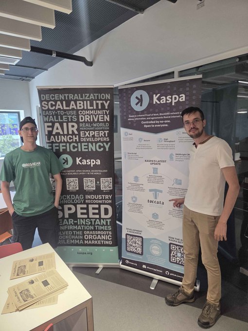 Two men pose in front of large "Kaspa" promotional banners at Imperial College London, with a table in the foreground displaying stacks of informational brochures and merchandise. The banners highlight key themes like "Decentralization," "Scalability," "Speed," and "Fair Launch," promoting $KAS as a "fastest, open-source, decentralized Layer 1" blockchain network.