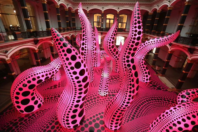A large-scale pink and black polka-dotted inflatable sculpture by Yayoi Kusama displayed in the ornate, multi-level interior of a museum.