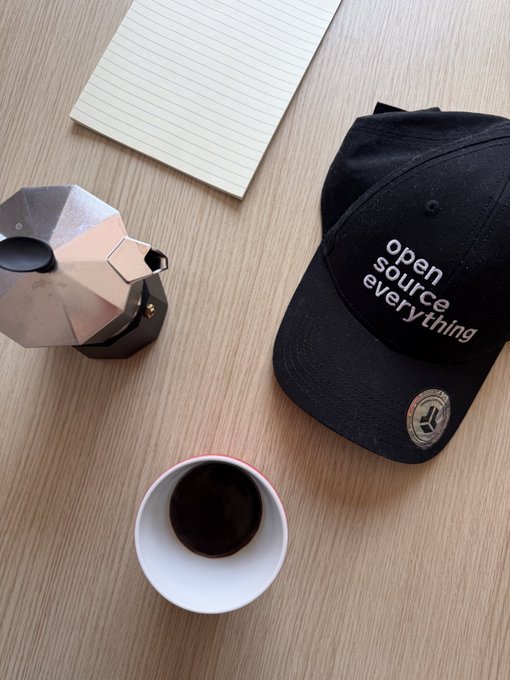 A flat-lay photo on a wooden desk featuring a black 'open source everything' baseball cap, an octagonal Moka pot coffee maker, a white cup of black coffee, and a lined notepad.