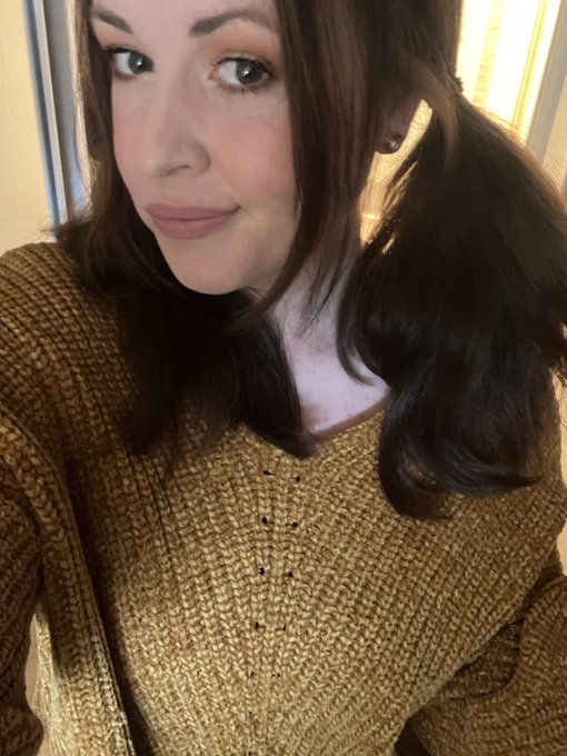A close-up selfie of a woman with dark brown hair in a ponytail, wearing a mustard-yellow knit sweater indoors.