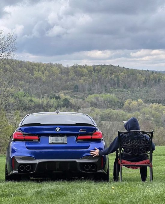 A person in a hoodie sits in a chair next to a blue BMW M5 parked on a grassy hill overlooking a forested valley.