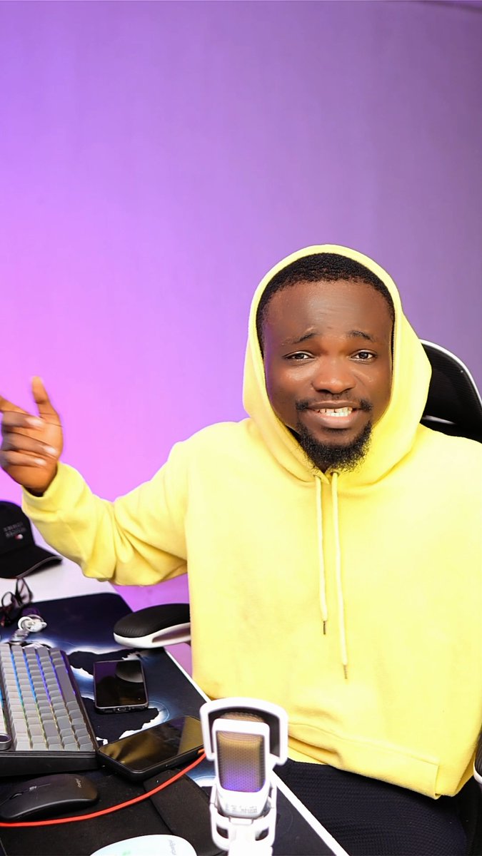A man wearing a yellow hoodie sitting in a gaming chair against a purple background, gesturing with his right hand behind a desk with a keyboard, phone, and microphone.