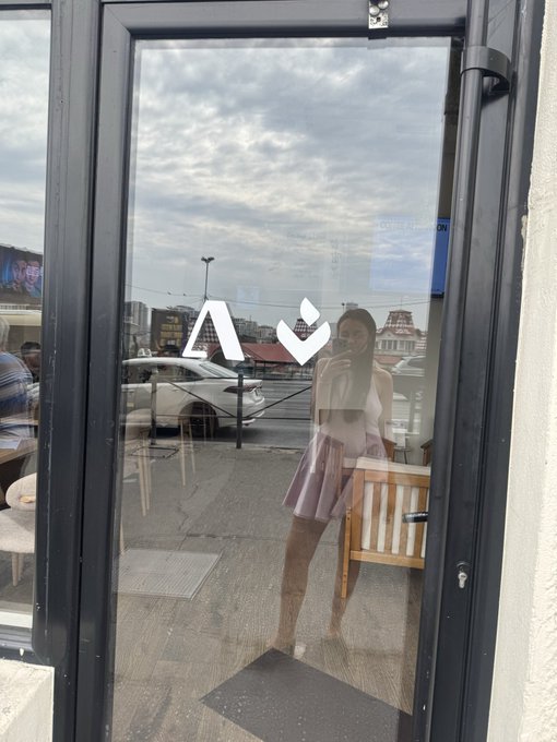 A reflection in a glass door shows a woman in a pink dress taking a mirror selfie, with a car and buildings visible in the background.