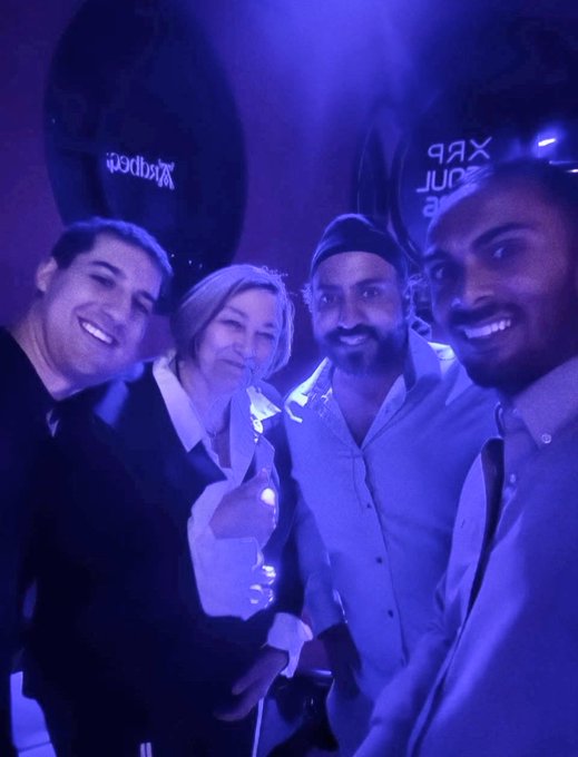 Four people pose together for a selfie at what appears to be a vibrant, neon-lit nightclub, with the second person flashing a peace sign. The blue/purple lighting matches the energetic atmosphere suggested by the post about an IRL meetup with "Panos, Amrit & Adam" at an @axelar and @Yellow event during @XRPSEOUL.