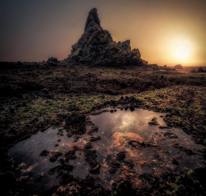 A dramatic landscape photograph captures a tall, jagged rock formation silhouetted against a sunset sky, with a tidal pool in the foreground reflecting the warm golden light.