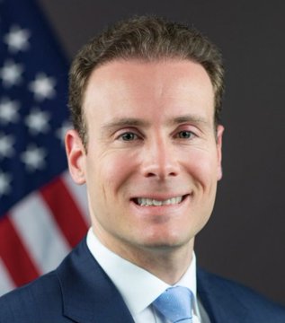 A professional headshot shows a smiling man with short brown hair, wearing a dark blue suit and light blue tie, posed in front of an American flag — identified as CFTC Chair Mike Selig in the accompanying post, where he is quoted saying the agency is "lock in and ready to ship its rules for the new frontier of finance" after the Senate failed to advance related legislation.