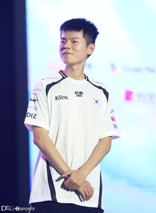 A young man wearing a white jersey with "KSPA," "R08L STUDIO," and "DIZ" logos stands with his hands clasped, smiling slightly, as a South Korean flag patch is visible on his sleeve—matching the post's welcome-back tribute to Deft, the League of Legends pro player.
