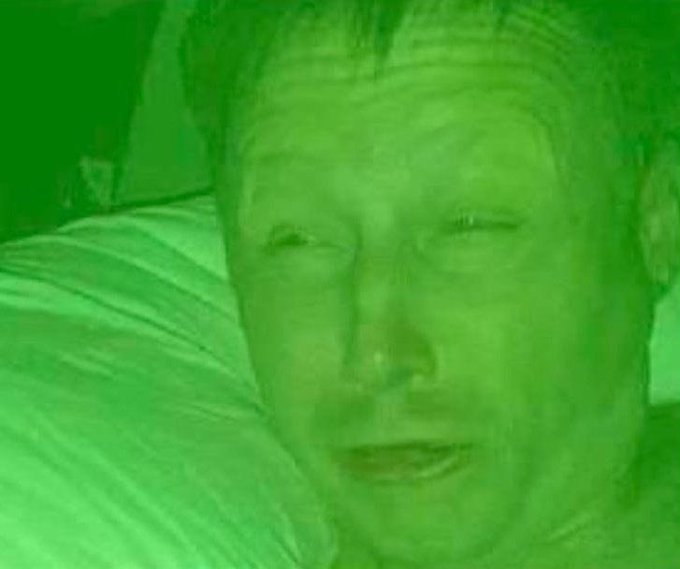 A close-up, green-tinted photo of a man lying in bed, appearing distressed or emotional, with the image heavily filtered in a monochromatic green hue.