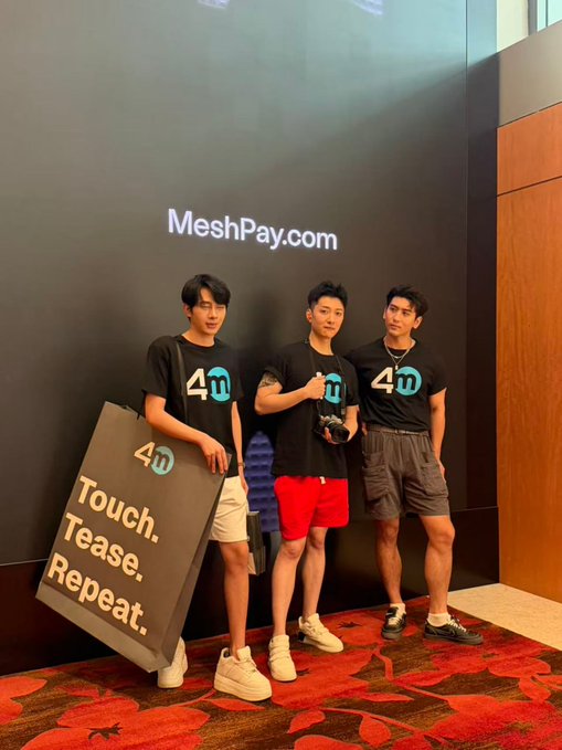 Three young men pose in matching black t-shirts featuring a '4m' logo, standing next to a 'MeshPay' visible text sign and a placard reading '4m Touch. Tease. Repeat.'